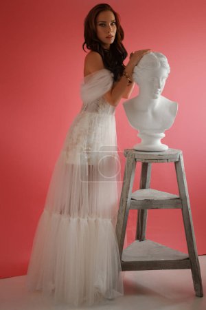 Foto de Hermosa morena en un delicado vestido blanco posando junto a un antiguo busto griego sobre un fondo rosa - Imagen libre de derechos