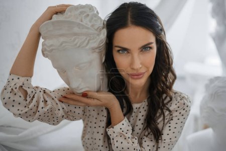 Photo for Brunette in dress holds ancient Greek sculpture head, posing amidst sculptors timeless art - Royalty Free Image