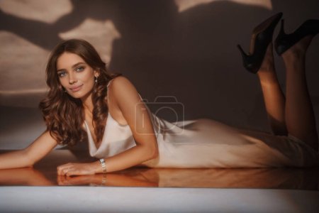 Téléchargez les photos : Femme enchanteresse en robe de soirée pose sur une surface miroir, coulant des reflets dorés dans tout le studio - en image libre de droit