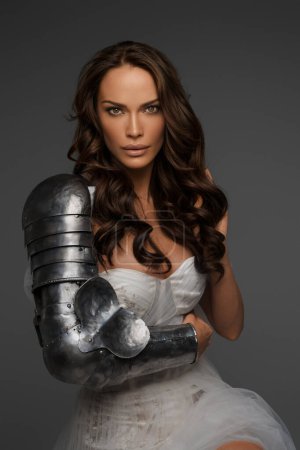 Photo for Elegant middle-aged brunette in a white gown and partial medieval armor poses on a gray backdrop - Royalty Free Image