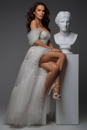 Foto de Mujer elegante en vestido blanco prístino de pie con gracia al lado de estatuas griegas antiguas - Imagen libre de derechos