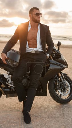 Photo for Elegantly dressed muscular man leans on his black sports bike on deserted beach at sunset - Royalty Free Image