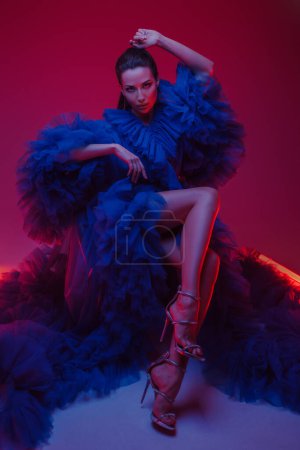 Téléchargez les photos : Beau modèle dans une somptueuse robe de carnaval bleue pose dans un studio chaleureusement éclairé, accentué par des néons - en image libre de droit