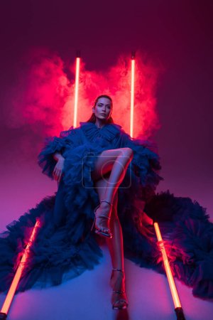 Beautiful model in a lavish blue carnival gown poses in a warmly lit studio, accentuated by neon lights
