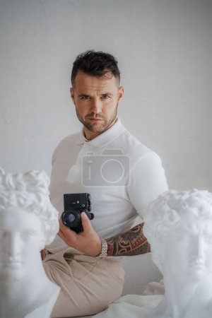 Foto de Hombre rico en una camisa blanca luciendo un tatuaje, posa con una cámara entre esculturas griegas antiguas en una habitación suavemente iluminada - Imagen libre de derechos