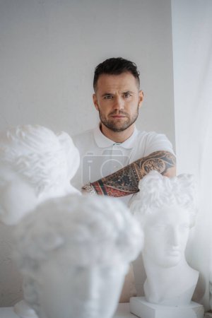 Foto de Hombre bien vestido con una camisa blanca con un tatuaje en el brazo posa en medio de antiguas esculturas griegas en una habitación suavemente iluminada - Imagen libre de derechos