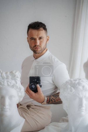 Foto de Hombre rico en una camisa blanca luciendo un tatuaje, posa con una cámara entre esculturas griegas antiguas en una habitación suavemente iluminada - Imagen libre de derechos