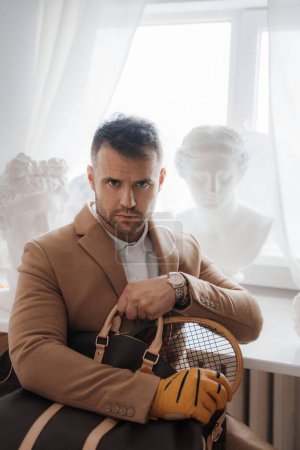 Téléchargez les photos : Homme riche dans un vieux style d'argent tenant un sac et une raquette de tennis dans un atelier de sculpteurs - en image libre de droit