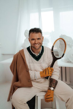Foto de Encantador hombre con estilo de dinero viejo, luciendo una sonrisa cautivadora, posa con una raqueta de tenis en medio de esculturas griegas antiguas - Imagen libre de derechos