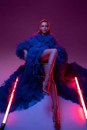 Téléchargez les photos : Un superbe modèle dans une somptueuse robe de carnaval bleu posant élégamment par la lueur chaude des néons dans un studio. - en image libre de droit