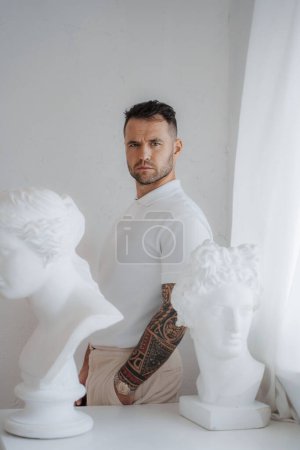 Téléchargez les photos : Homme bien habillé dans une chemise blanche avec un tatouage sur le bras pose au milieu des sculptures grecques antiques dans une pièce doucement éclairée - en image libre de droit