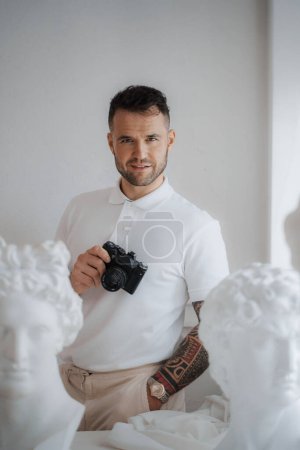 Photo for Wealthy man in a white shirt sporting a tattoo, poses with a camera among ancient Greek sculptures in a softly lit room - Royalty Free Image