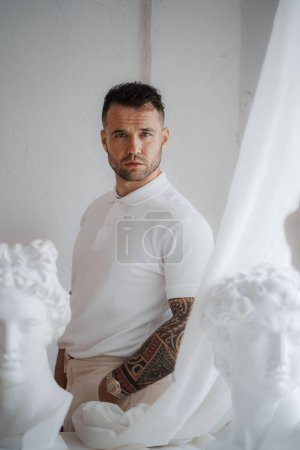 Foto de Hombre bien vestido con una camisa blanca con un tatuaje en el brazo posa en medio de antiguas esculturas griegas en una habitación suavemente iluminada - Imagen libre de derechos