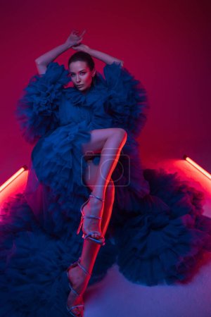 Foto de Un modelo impresionante en un lujoso vestido de carnaval azul posando elegantemente por el cálido brillo de las luces de neón en un estudio. - Imagen libre de derechos