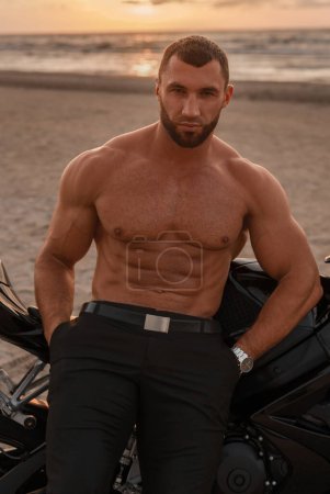 Rugged man with shades and a toned torso stands beside his black sports motorcycle on a desolate beach, with a cloudy sunset backdrop