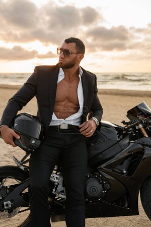 Foto de Hombre con estilo en un traje negro muestra su pecho musculoso, posando con su motocicleta contra una puesta de sol vívida en una playa - Imagen libre de derechos