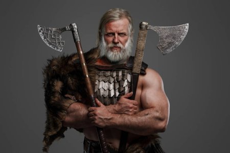 Foto de Héroe vikingo envejecido y venerable, vestido de piel y armadura ligera, luciendo un casco montado en el cinturón, agarrando dos ejes contra un telón de fondo neutral - Imagen libre de derechos