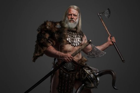 Foto de Anciano guerrero vikingo canoso, mostrando fuerza y sabiduría, vestido con pieles y armadura ligera, con un casco colgado en la cintura, blandiendo hachas dobles sobre un fondo neutro - Imagen libre de derechos