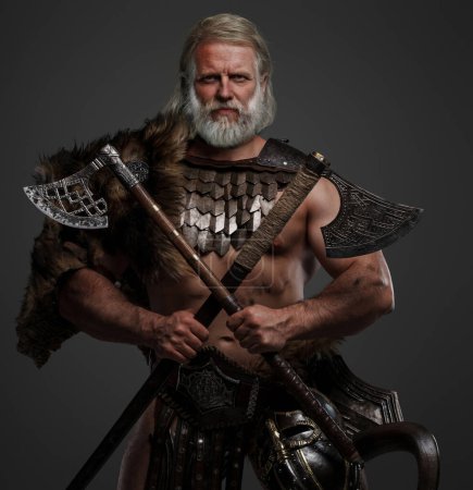 Foto de Héroe vikingo envejecido y venerable, vestido de piel y armadura ligera, luciendo un casco montado en el cinturón, agarrando dos ejes contra un telón de fondo neutral - Imagen libre de derechos