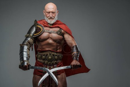 Foto de Gladiador barbudo maduro con cabeza calva, con armadura ligera y capa roja, empuñando dos espadas y de pie ante un fondo gris - Imagen libre de derechos