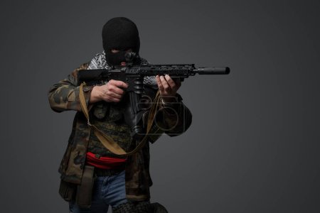 Foto de Retrato de un militante de Oriente Medio, con un traje de pasamontañas y camuflaje negro, sosteniendo un rifle, montado sobre un fondo gris - Imagen libre de derechos