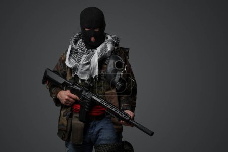 Téléchargez les photos : Soldat radical du Moyen-Orient vêtu d'une cagoule noire et d'un uniforme de campagne camouflé, armé d'un fusil, sur fond gris - en image libre de droit