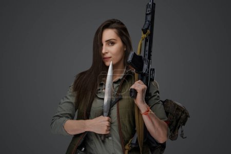 Foto de Mujer de Oriente Medio, vestida con equipo post-apocalíptico de supervivencia, posa con una daga y un rifle de asalto contra un fondo gris, encarnando la resiliencia y la preparación - Imagen libre de derechos
