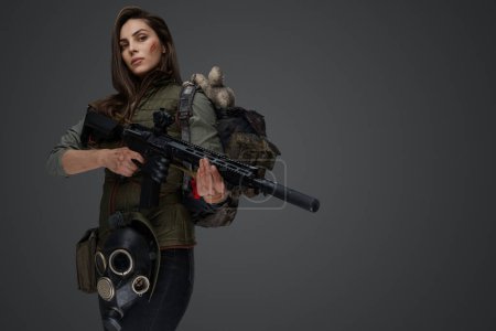 Foto de Mujer de aspecto de Oriente Medio vestida con ropa de supervivencia posando con un rifle sobre un fondo gris, retratando la fuerza y la resiliencia - Imagen libre de derechos