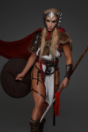 Photo for A muscular blonde Viking woman in fantasy armor with a red cloak, baring her thighs, poses holding a spear and shield against a gray background - Royalty Free Image