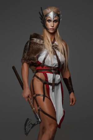 Photo for A muscular blonde Viking woman in fantasy armor poses holding two axes against a gray background - Royalty Free Image
