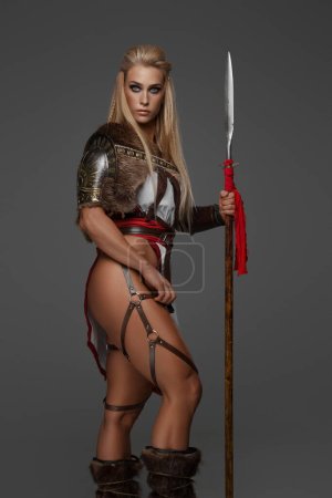 Foto de Una mujer vikinga rubia musculosa con armadura de fantasía posa sosteniendo una lanza sobre un fondo gris - Imagen libre de derechos