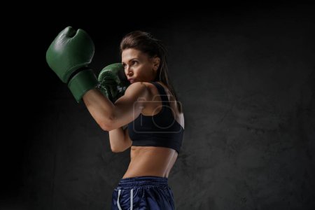 Photo for Athletic female fighter in sports bra, shorts, and boxing gloves showcases striking techniques against a dark backdrop - Royalty Free Image