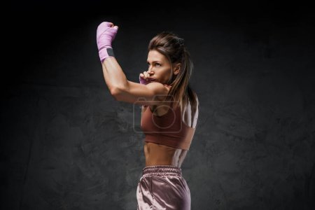 Téléchargez les photos : Boxeuse musclée démontrant des compétences frappantes, vêtue de vêtements de sport, dans un contexte sombre et lunatique - en image libre de droit