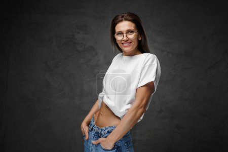 Photo for Radiant female in glasses and a white cropped tee proudly displays her toned core on a dark setting - Royalty Free Image