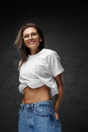 Photo for Gleaming woman in glasses and a white crop-top unveils her chiseled abs against a dark backdrop - Royalty Free Image