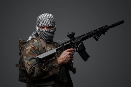 Foto de Militante de Oriente Medio, vestido con un keffiyeh blanco y un traje camuflado de campo, con un rifle automático, montado sobre un fondo gris neutro - Imagen libre de derechos