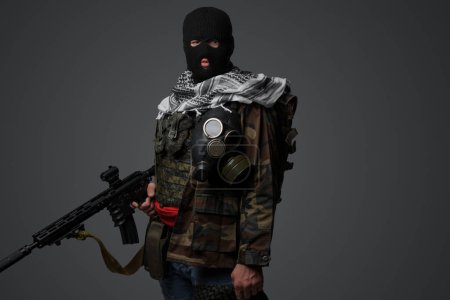 Photo for A Middle Eastern radical soldier dressed in a black balaclava and camouflaged field uniform, armed with a rifle, against a gray background - Royalty Free Image