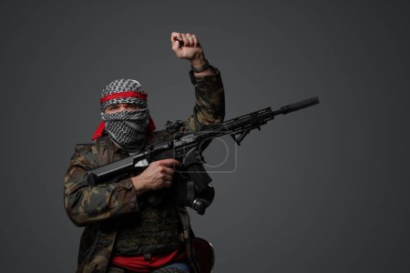 Photo for Fanatic soldier from the Middle East, wearing a white keffiyeh and camouflaged field uniform, armed with a rifle, posing with a raised hand against a gray background - Royalty Free Image