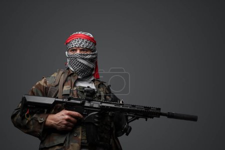Photo for Militant from the Middle East, dressed in a white keffiyeh and camouflaged field attire, wielding an automatic rifle, set against a neutral gray backdrop - Royalty Free Image
