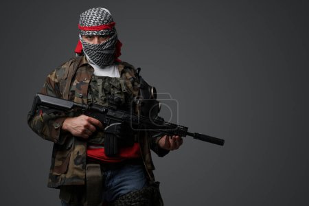Foto de Militante de Oriente Medio, vestido con un keffiyeh blanco y un traje camuflado de campo, con un rifle automático, montado sobre un fondo gris neutro - Imagen libre de derechos
