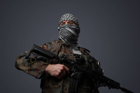 Téléchargez les photos : Soldat fanatique radical du Moyen-Orient revêtu d'un keffiyeh blanc et d'un uniforme de campagne camouflé, armé d'un fusil automatique, sur fond gris - en image libre de droit