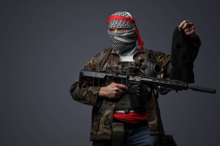 Foto de Militante de Oriente Medio, vestido con un keffiyeh blanco y camuflado traje de campo, empuñando un rifle, posando con un pasamontañas negro sostenido en la mano contra un fondo gris - Imagen libre de derechos