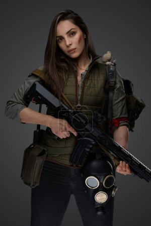 Téléchargez les photos : Femme d'apparence moyen-orientale vêtue de vêtements de survie posant avec un fusil sur un fond gris, dépeignant la force et la résilience - en image libre de droit