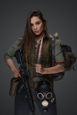 Foto de Mujer de aspecto de Oriente Medio vestida con ropa de supervivencia posando con un rifle sobre un fondo gris, retratando la fuerza y la resiliencia - Imagen libre de derechos