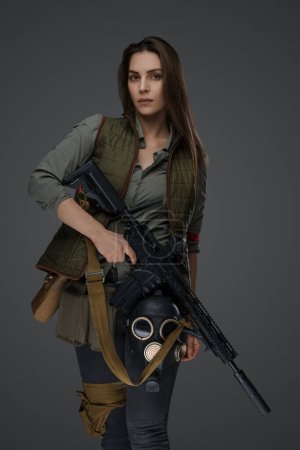 Photo for Middle Eastern-looking woman dressed in survivalist clothing posing with a rifle against a gray background, portraying strength and resilience - Royalty Free Image