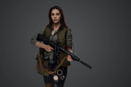 Photo for Middle Eastern-looking woman dressed in survivalist clothing posing with a rifle against a gray background, portraying strength and resilience - Royalty Free Image