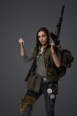 Foto de Mujer de Oriente Medio, vestida con equipo post-apocalíptico de supervivencia, posa con una daga y un rifle de asalto contra un fondo gris, encarnando la resiliencia y la preparación - Imagen libre de derechos