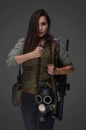 Photo for Middle Eastern descent in post-apocalyptic survival gear, brandishing a dagger on a gray backdrop, showcasing her determination to survive - Royalty Free Image