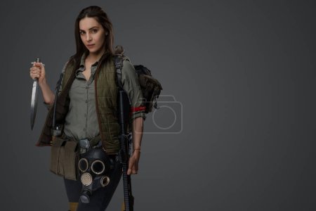 Photo for Middle Eastern descent in post-apocalyptic survival gear, brandishing a dagger on a gray backdrop, showcasing her determination to survive - Royalty Free Image