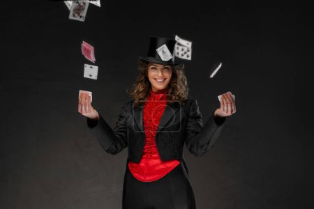 Foto de Encantadora mago femenino en un disfraz de magos y sombrero de copa negro realizar trucos de cartas sobre un fondo oscuro - Imagen libre de derechos